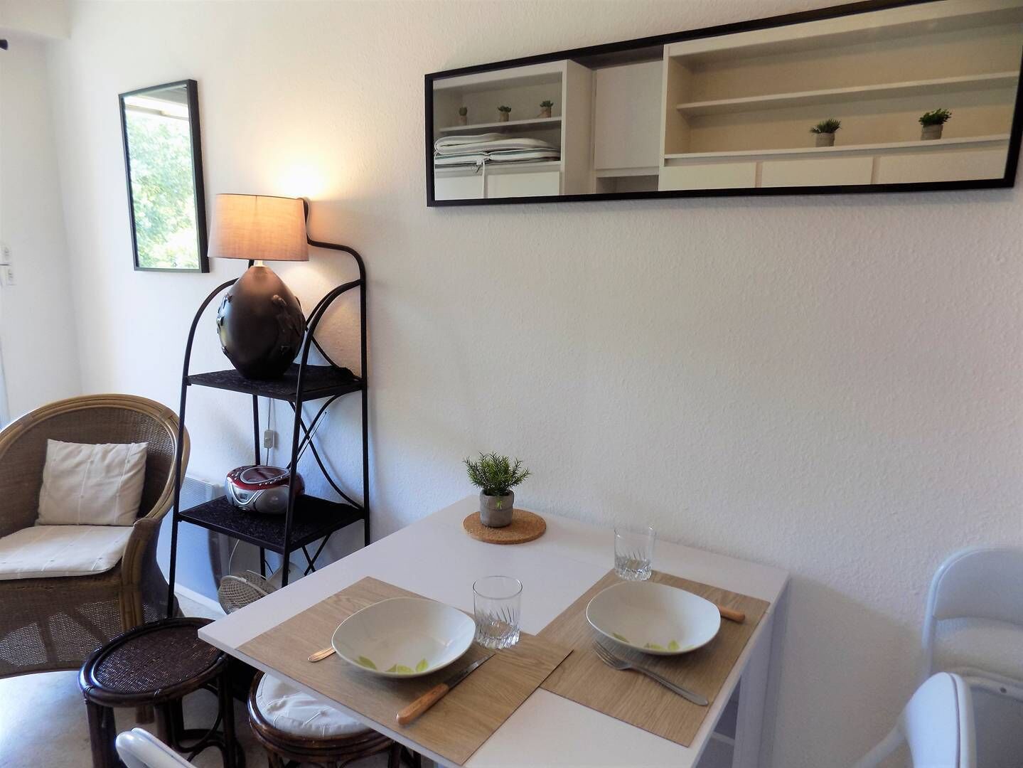 A dining area with a table set for two, a wicker chair, a lamp, and a shelf with plants.