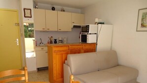 Une cuisine compacte équipée d’un four à micro-ondes, d’une machine à café et d’un évier. On y trouve également un canapé beige et une table en bois avec des chaises.