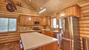 Cocina de cabaña de troncos con armarios de madera, un refrigerador de acero inoxidable, un microondas y una gran cabeza de alce montada en la pared.