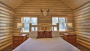 Un dormitorio tipo cabaña de madera con cama, dos mesitas de noche, lámparas y un gran cráneo de toro en el techo.