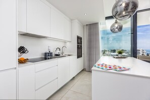 A modern kitchen with white cabinetry, a built-in oven, and a large window overlooking a cityscape. - Ivy | Sea view - bbq - terrace (Fuengirola)