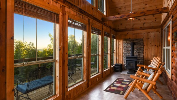 A log cabin interior with large windows, a stone fireplace, and a deck with outdoor furniture.