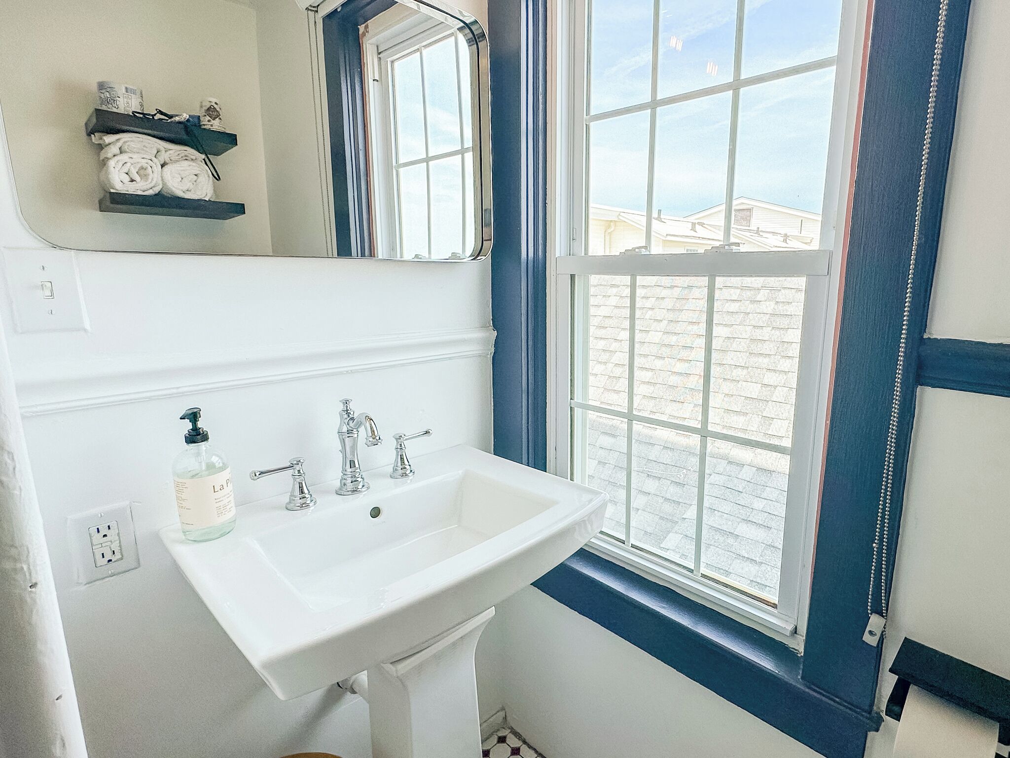 Une salle de bain avec un lavabo blanc, un miroir et une fenêtre donnant sur les bâtiments environnants.