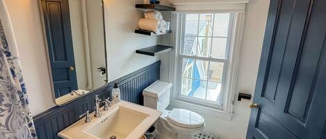 Un baño con lavamanos blanco, inodoro, espejo y ventana con vistas a edificios.