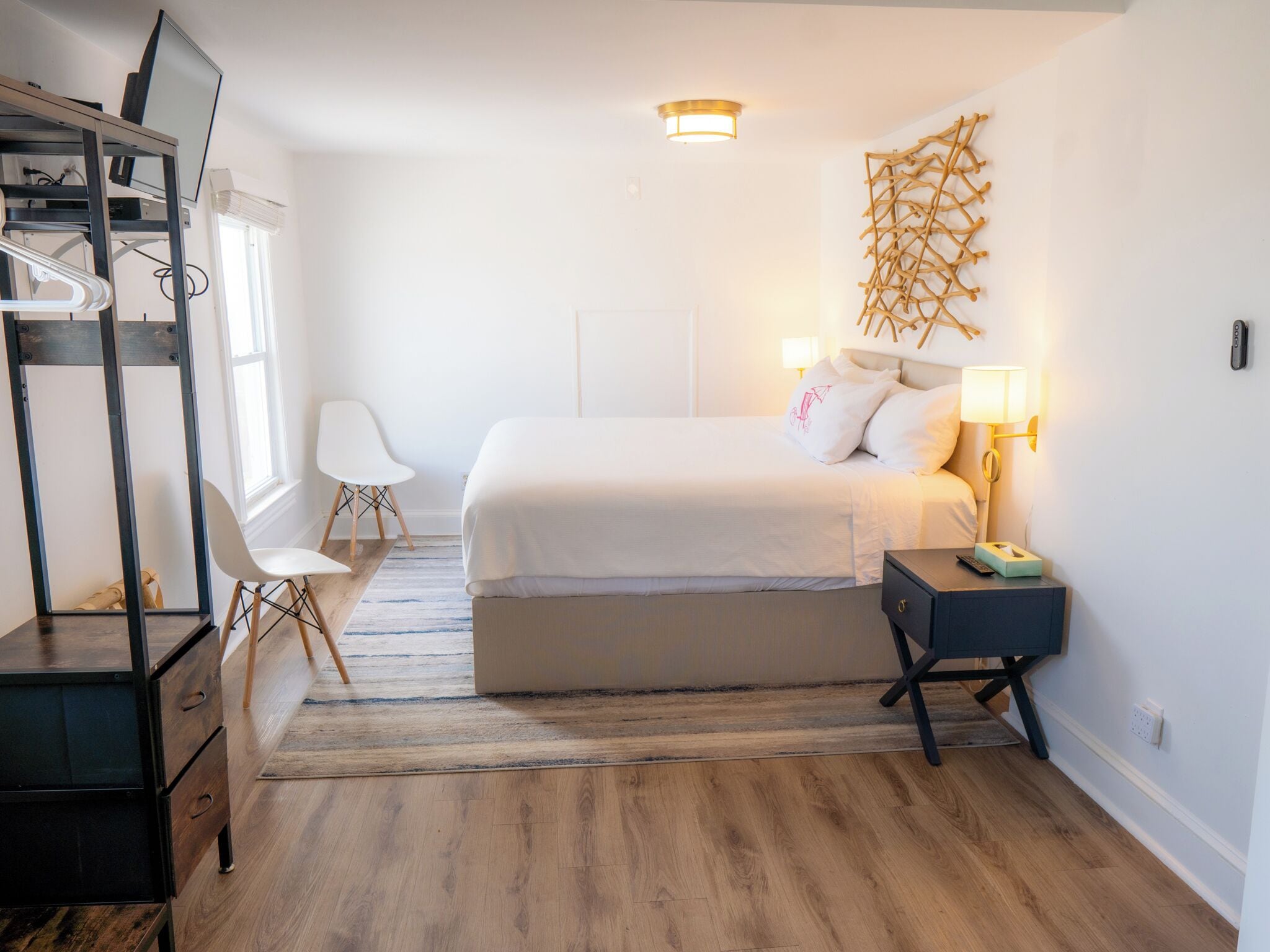 Una habitación de hotel moderna con cama, mesita de noche, lámpara, silla, escritorio y un elemento decorativo en la pared.