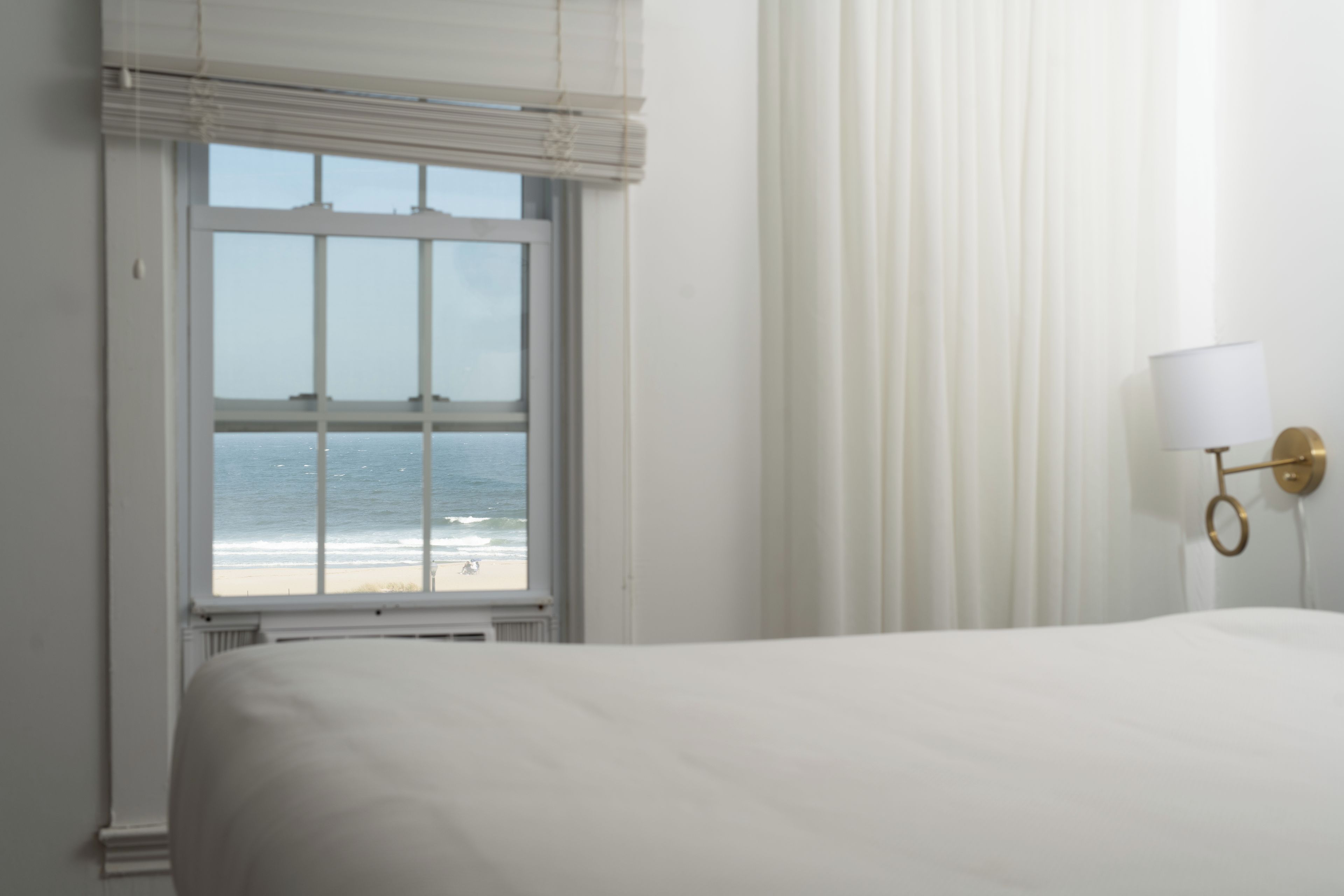A hotel room with a large bed, a window with a view of the beach, and a wall-mounted lamp.