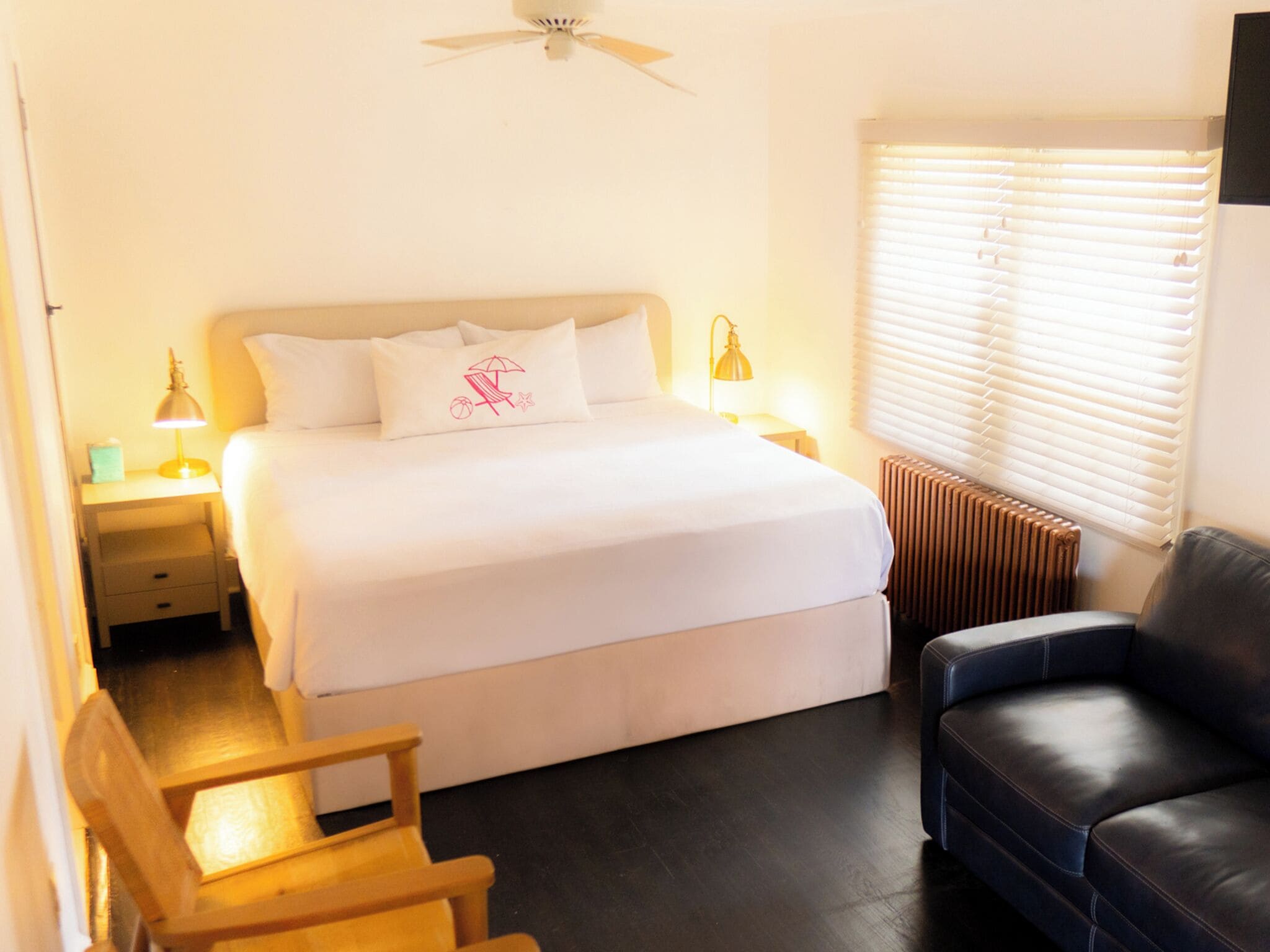 A bedroom with a bed, bedside lamps, a radiator, a window with blinds, a wooden chair, and a leather sofa.