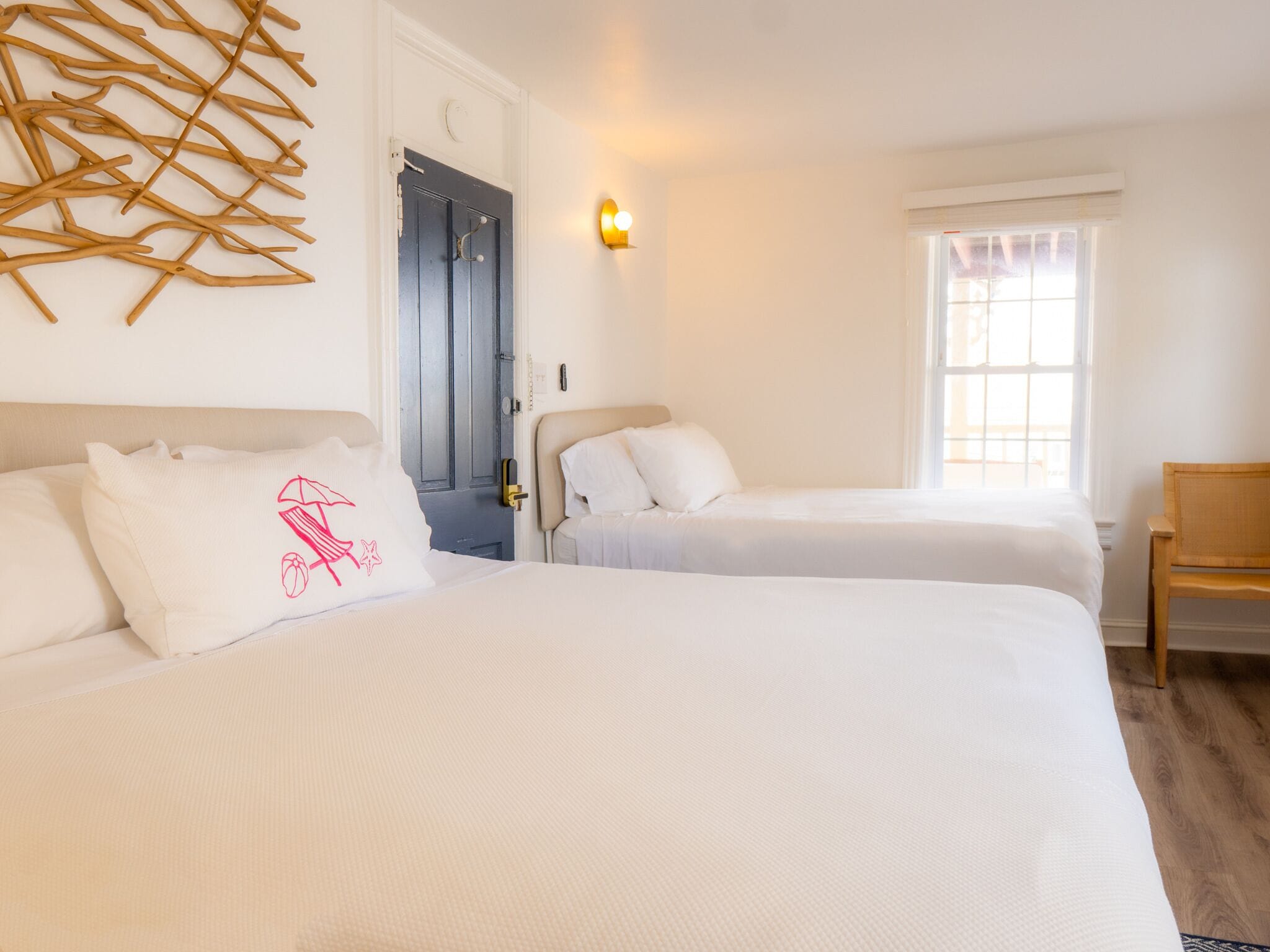 A hotel room with two beds, a wooden chair, a wall-mounted light, and a door with a handle.