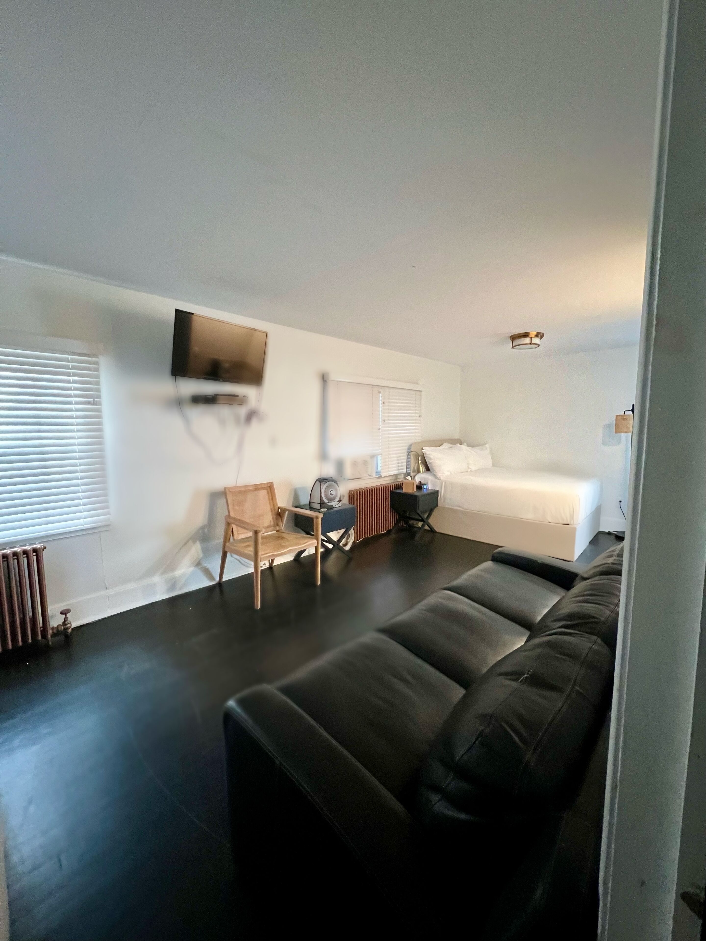 A modern, compact living space with a black leather sofa, a small wooden chair, a bed, and a television mounted on the wall.