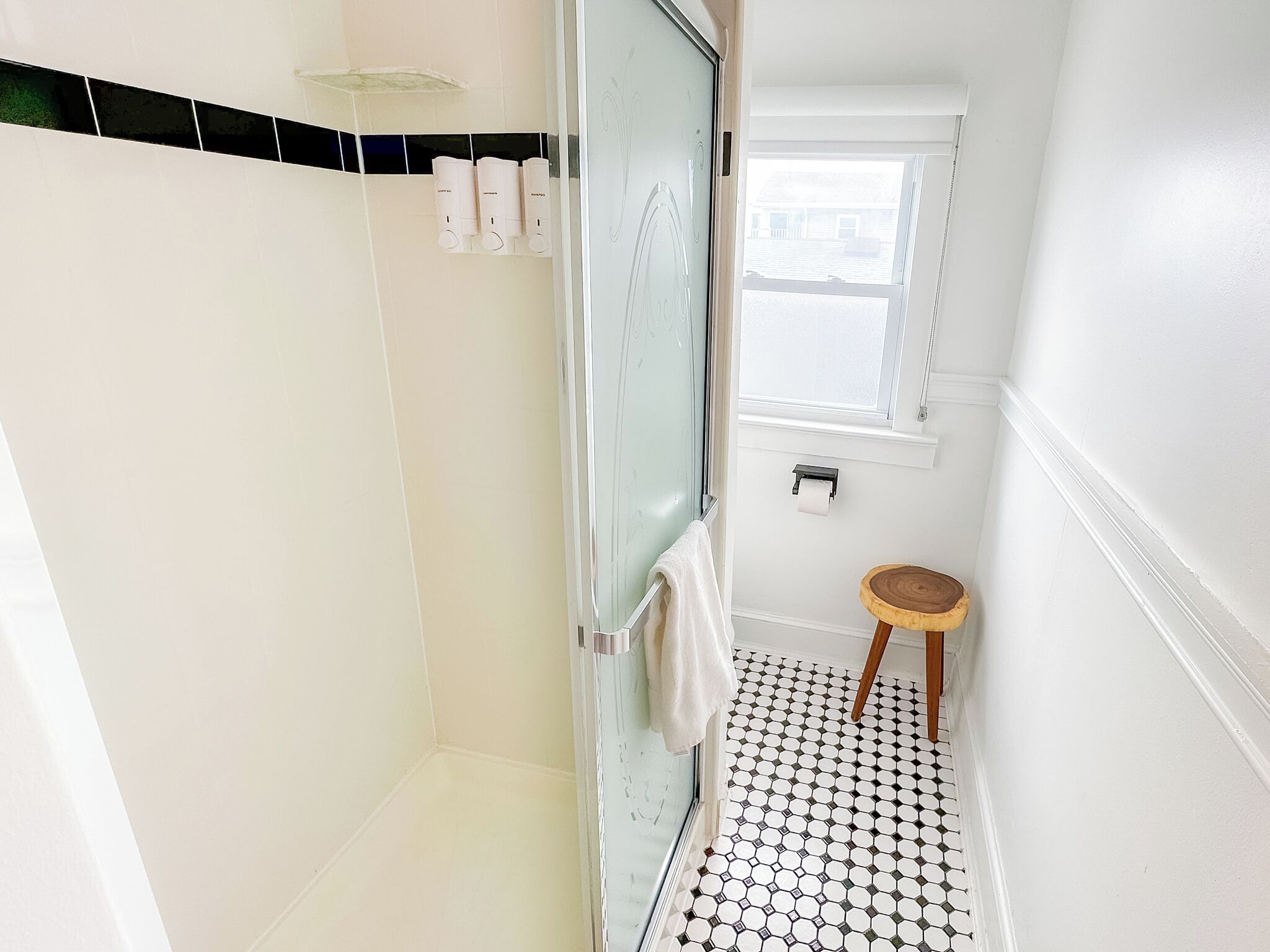 A bathroom with a glass shower enclosure, a toilet paper holder, a small wooden stool, and a window providing natural light.