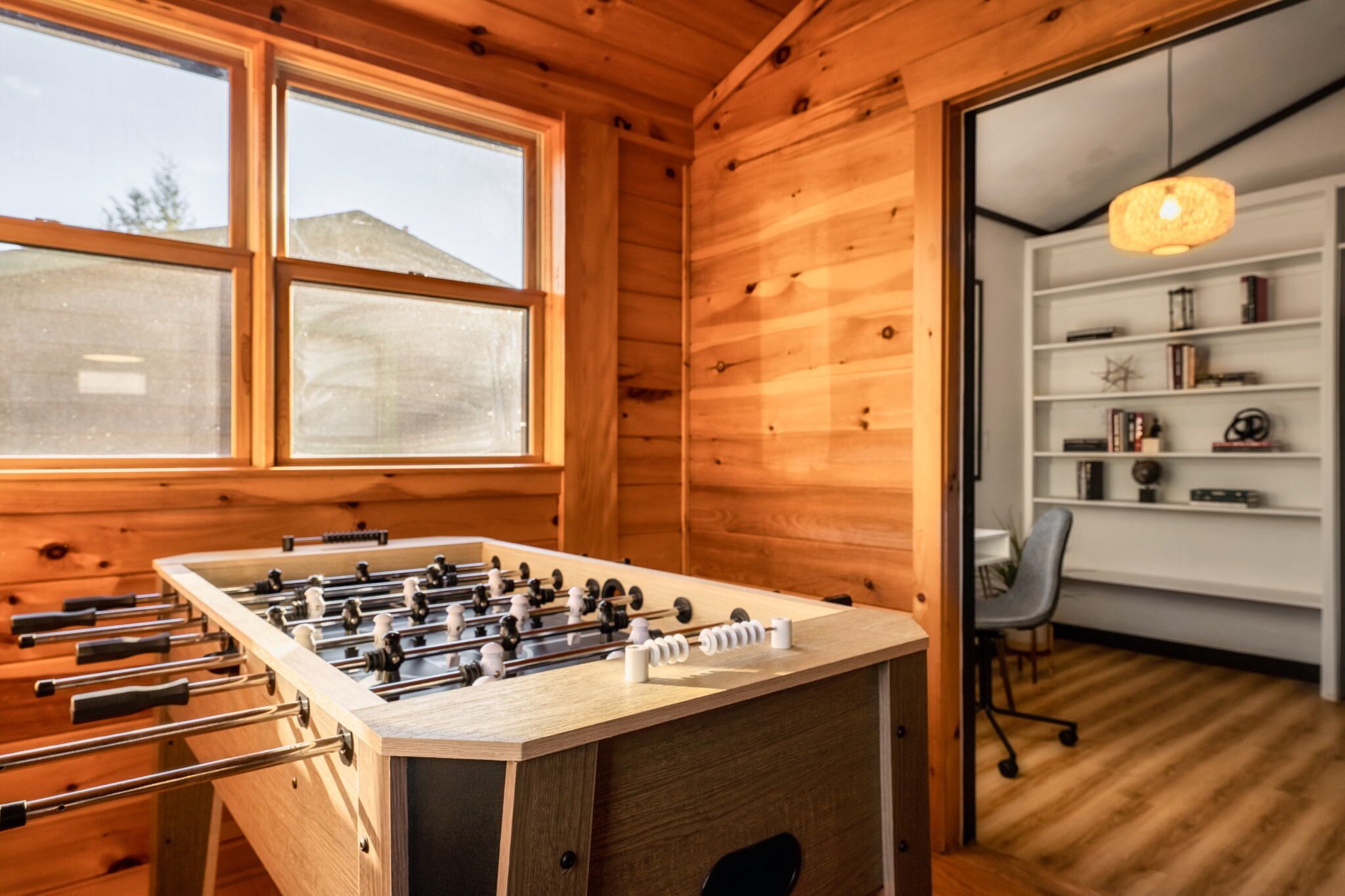A wooden-floored room with a foosball table, large windows, and a bookshelf with various items.