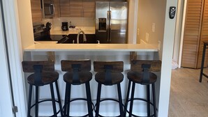 A compact kitchen with wooden cabinets, a stainless steel refrigerator, and a bar stool area. - BEAUTIFUL & SPACIOUS 2BR/2BA BEACH CONDO (Jacksonville Beach)