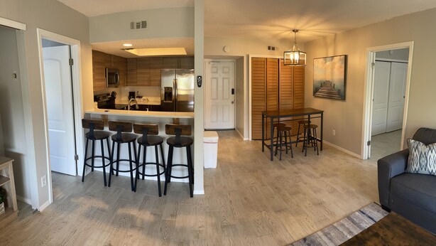A modern living room with a dark-colored sofa, a coffee table, and a dining area with a small table and four stools. A kitchen with a large island and bar stools is visible in the background. - BEAUTIFUL & SPACIOUS 2BR/2BA BEACH CONDO (Jacksonville Beach)