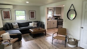 A modern living room with a dark grey sofa, a wooden coffee table, and a wicker chair. A kitchen is visible in the background with light wooden cabinets and a stove. - Detox & Hike: Waterfront Oasis with Infrared Sauna & Shawangunk Mountains (wallkill)