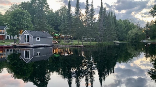 Eine Hütte an einem See, mit Spiegelung von Bäumen und einem Haus am anderen Ufer.