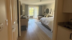 Quarto de hotel com cama, escrivaninha, cadeira, mesinha e cozinha compacta.
