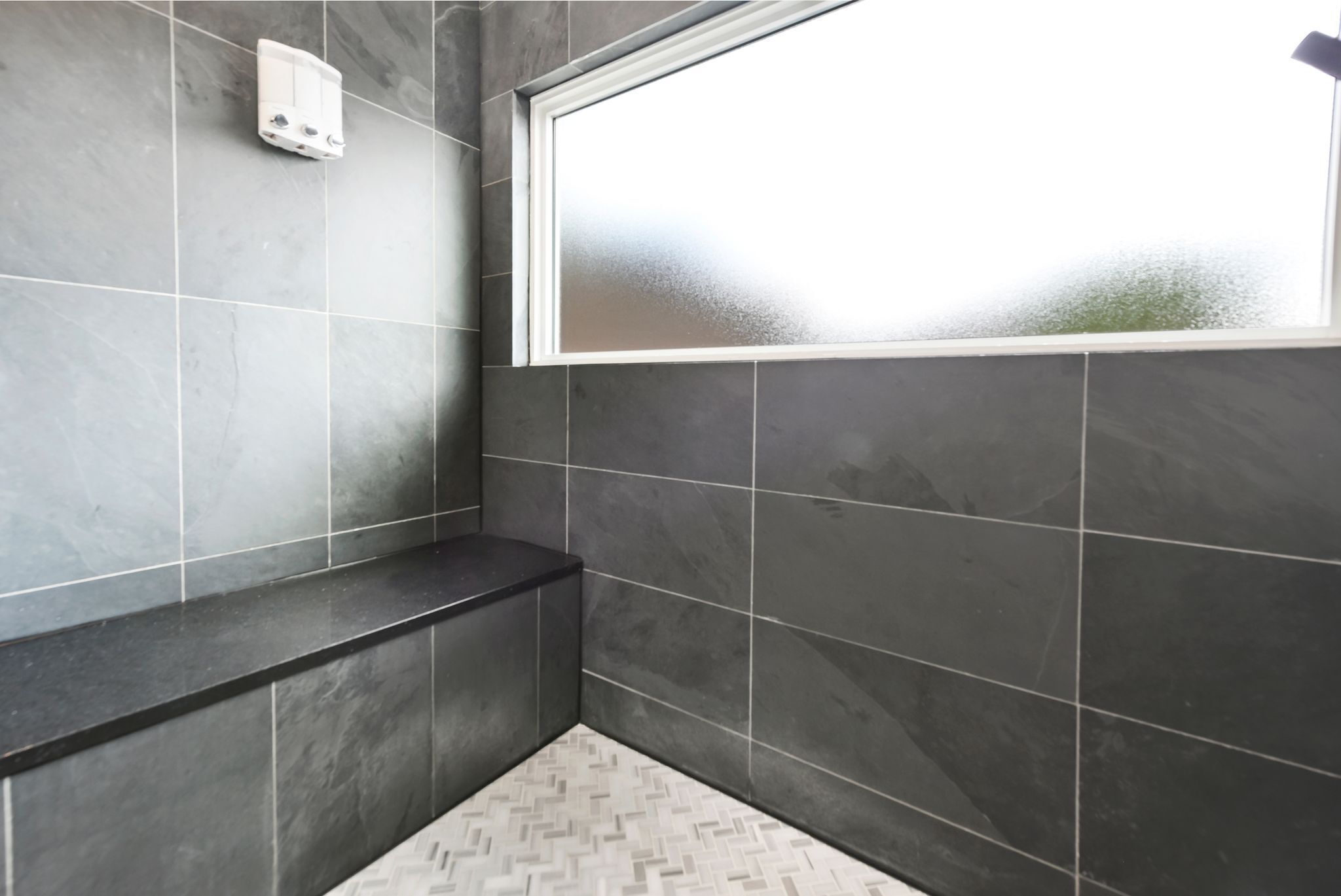 Une salle de bain avec des murs recouverts de carreaux foncés, une fenêtre blanche et un sol à motifs en chevrons.
