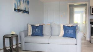 Un saló amb un sofà gris clar, dos coixins amb els textos "BEACH" i "HOUSE", una taula rodona petita i un quadre de velers.