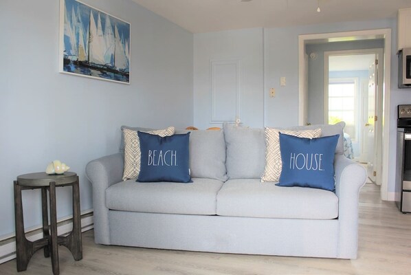 A living room with a light grey sofa, two pillows with "BEACH" and "HOUSE" text, a small round table, and a painting of sailboats.