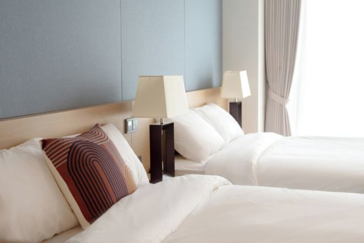 A hotel room with two beds, each with white linens and a single decorative pillow. There are bedside tables with lamps, and the room has a neutral color scheme with blue and beige walls.