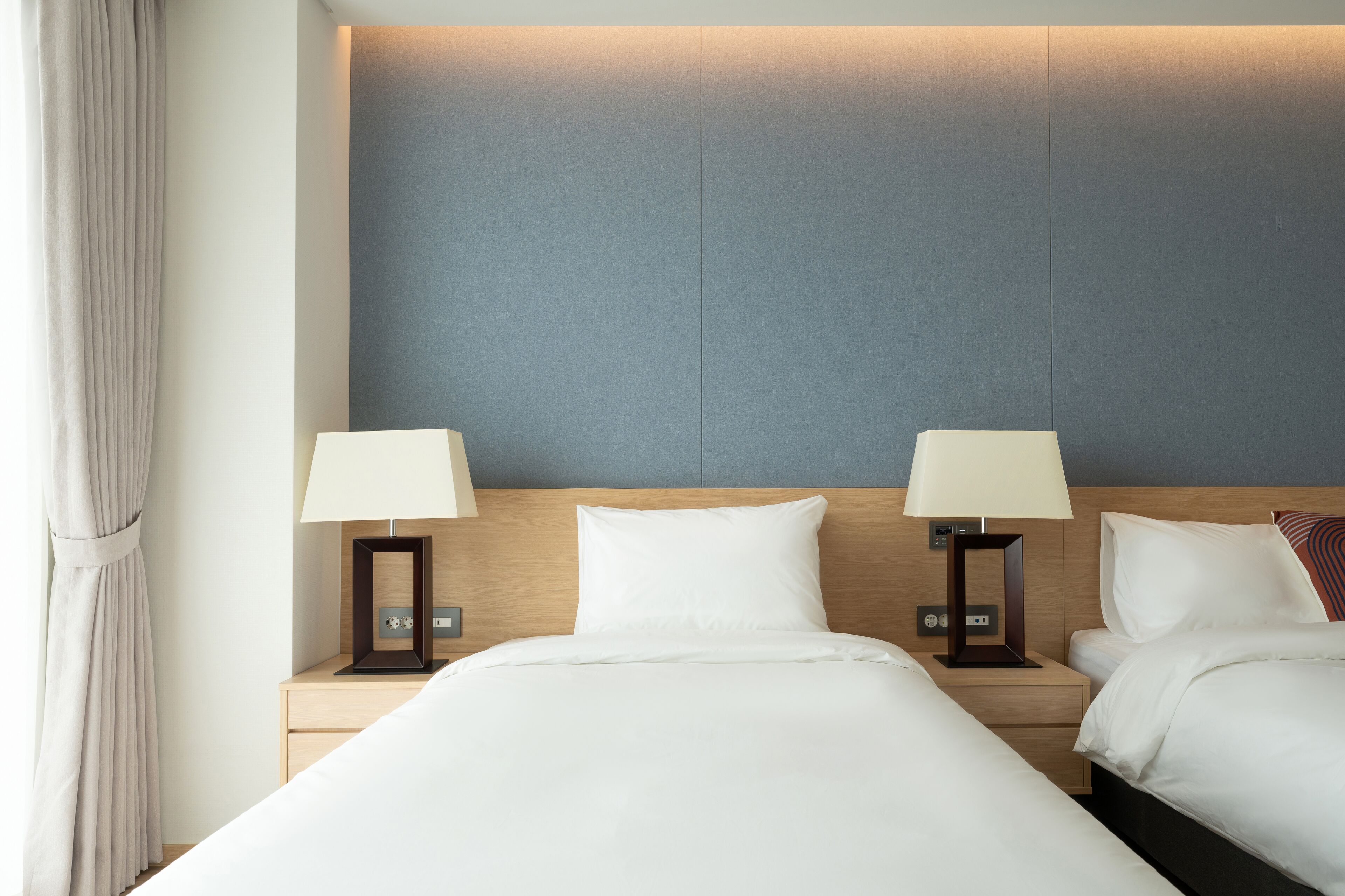 A hotel room with two beds, each with a bedside table and lamp. The room has a neutral color scheme with a blue accent wall.