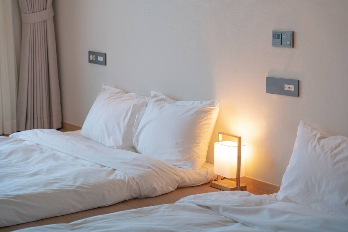 A neatly made bed with white bedding, a bedside lamp, and two wall-mounted switches.
