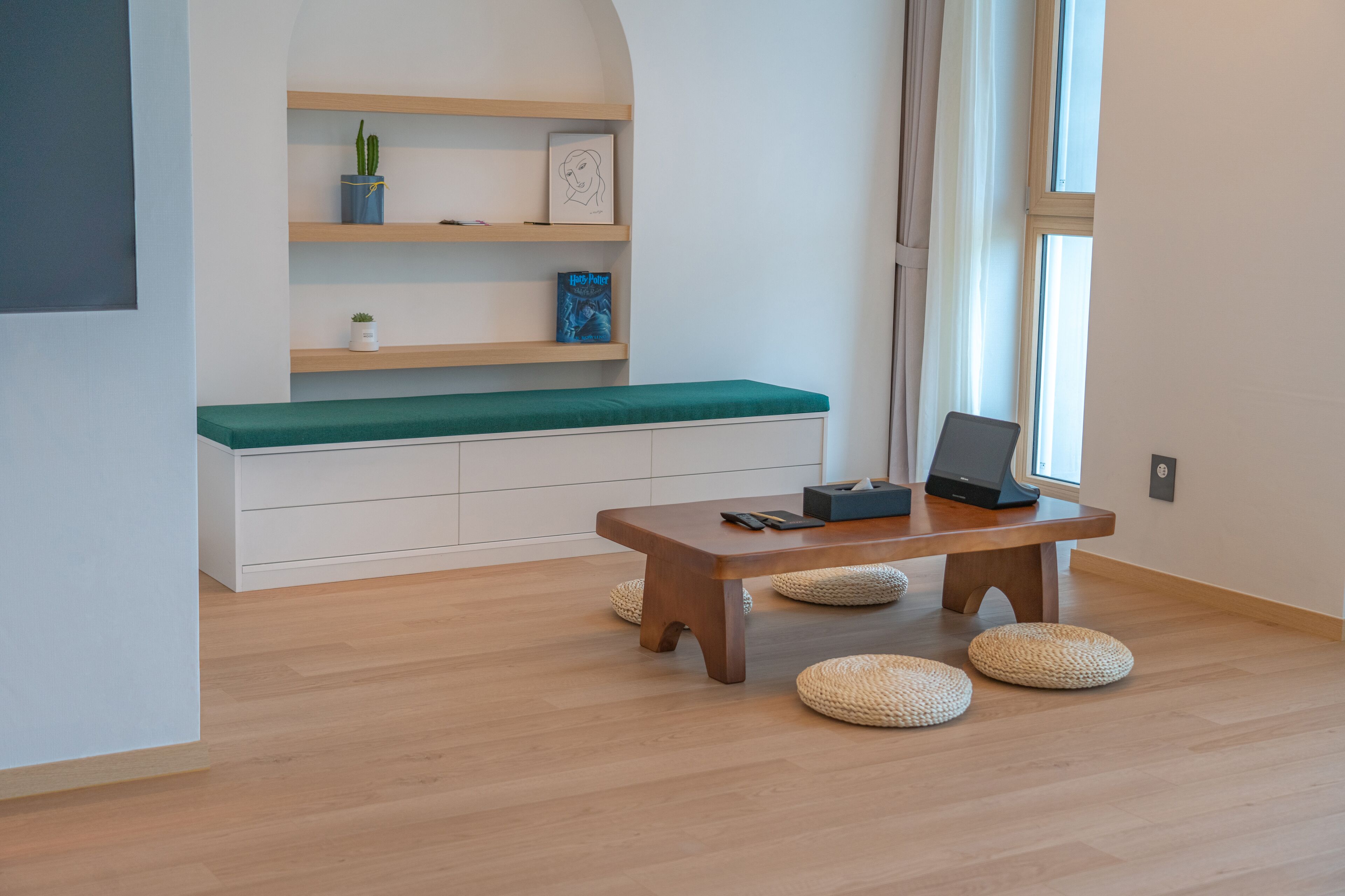 Una habitación moderna con piso de madera, un banco acolchado verde, una mesa de madera con una computadora portátil y pufs tejidos.