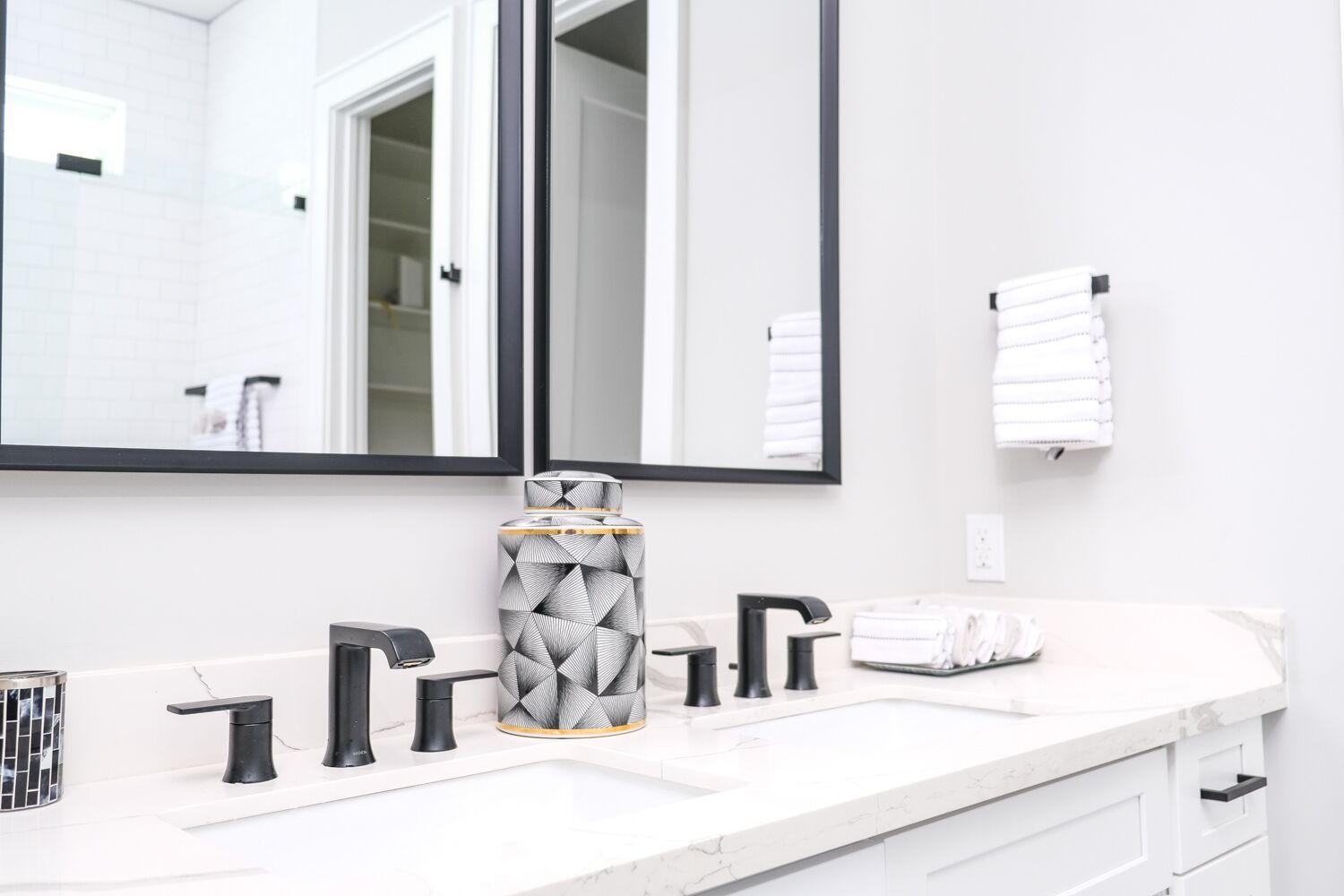 Casa de banho moderna com toucador branco, torneiras pretas, um espelho grande e um recipiente com padrão geométrico.