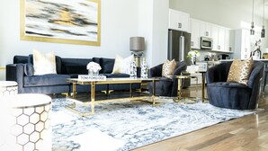 Un salon moderne avec un canapé bleu foncé, une table basse et un tapis. On aperçoit au fond une cuisine avec des meubles blancs et un réfrigérateur.