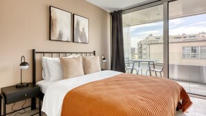 Habitación de hotel con cama, mesita de noche, escritorio y balcón con vistas a edificios.