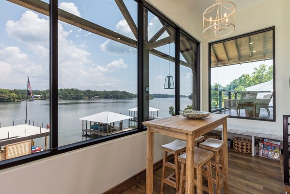 Una habitación con ventanas amplias que dan a un lago, una mesa de madera con sillas, y una lámpara colgante.