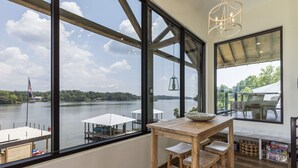 A room with large windows overlooking a lake, a wooden table with chairs, and a hanging light fixture.