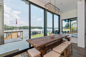 Un comedor espacioso con una gran mesa de madera y taburetes de bar, con vista a un lago con un muelle y sombrillas.