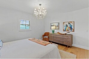 A bedroom with a bed, a dresser, a chair, a chandelier, and two paintings on the wall.