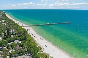En strand med en brygga som sträcker sig ut i havet, omgiven av bostadshus och grönska.