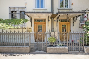 A building with two numbered doors, a wrought iron fence, and a small garden area with potted plants.