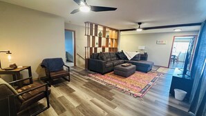 Ein Wohnzimmer mit einer Couch, Sesseln, einem Couchtisch, einem gemusterten Teppich, einem Deckenventilator und einer Treppe, die in einen anderen Raum führt.