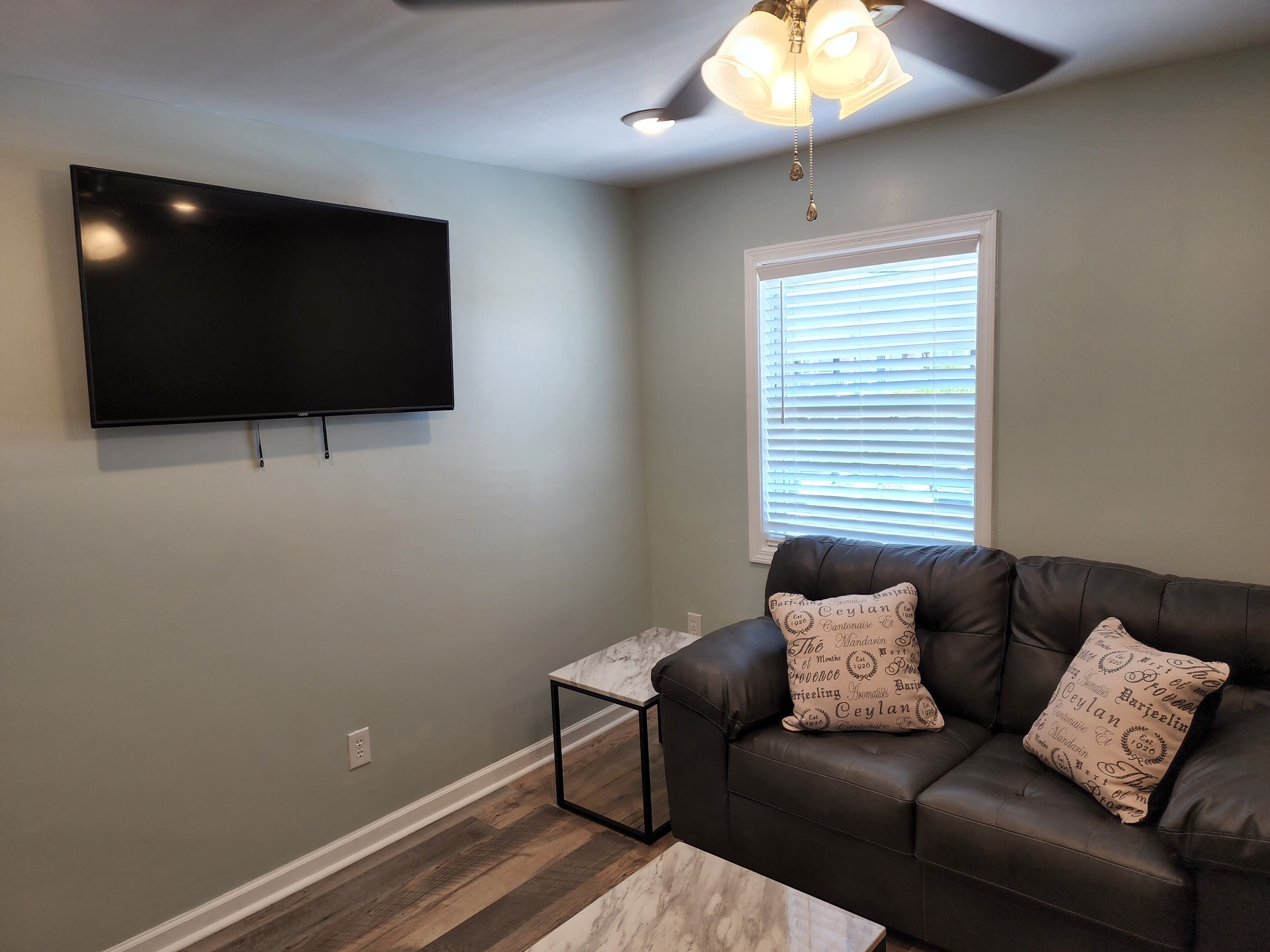 A living room with a flat-screen TV mounted on the wall, a leather sofa with patterned cushions, a small side table, and a window with blinds.