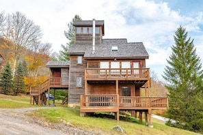 Une cabane en bois à deux étages, dotée d’un balcon et d’une cheminée, entourée d’arbres et bordée d’un chemin de gravier.