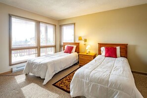 A hotel room with two beds, a nightstand with a lamp, and large windows with blinds.