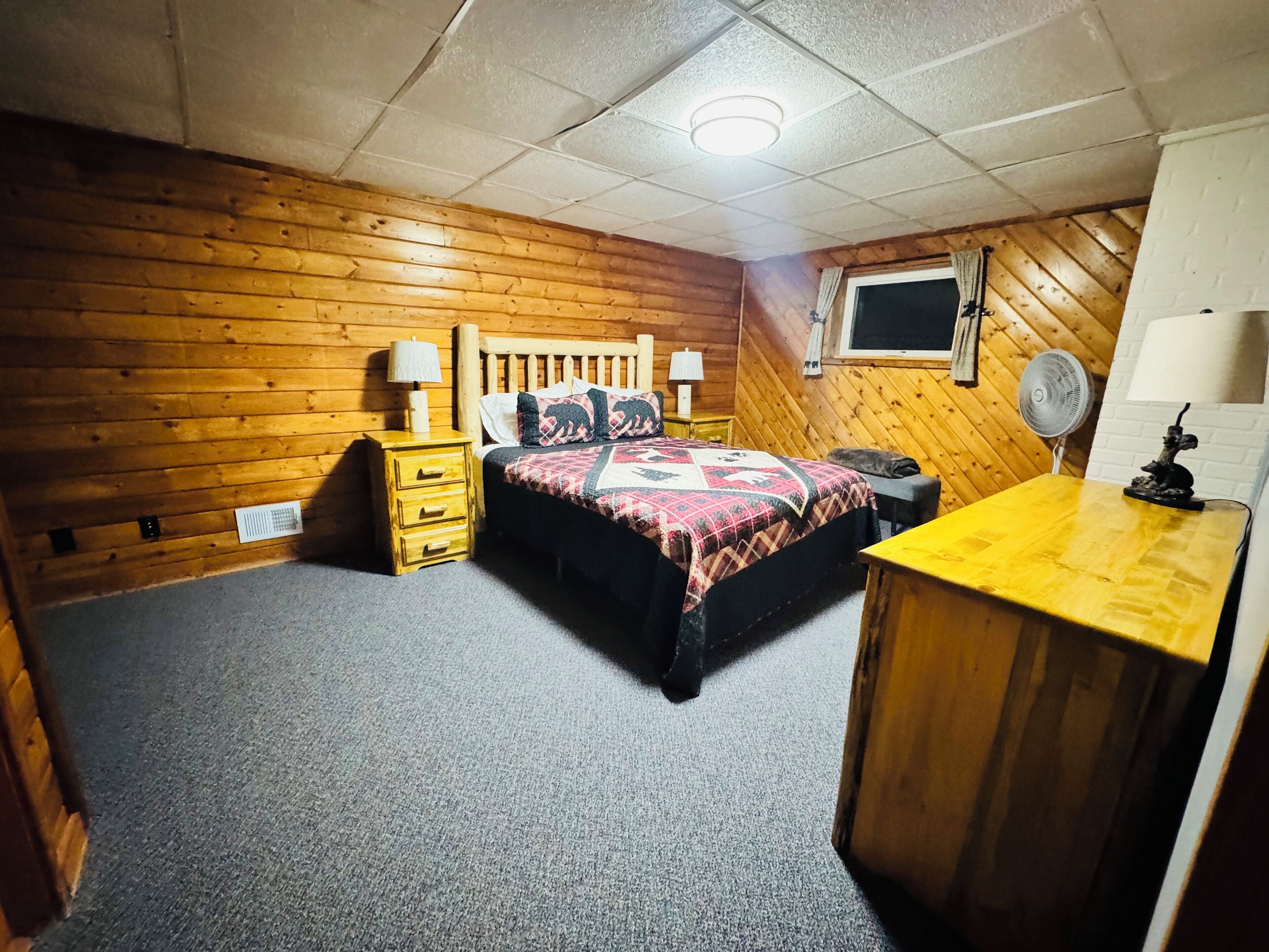 A bedroom with a wooden floor, a bed with a patterned blanket, two bedside tables with lamps, a small desk with a lamp, and a window with a view of the outside.