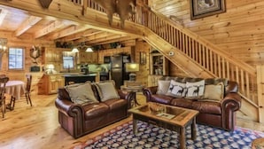 Interior de una cabaña de madera con escalera, sofás de cuero, zona de comedor y trofeos de animales colgados.