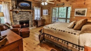 Una habitación tipo cabaña de madera con chimenea, una cama con ropa de cama estampada, un ventilador de techo y vistas al exterior.