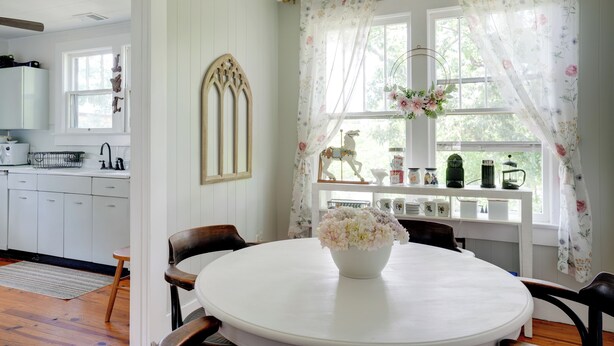A dining area with a round table, chairs, a vase of flowers, a window with floral curtains, and a view of the kitchen.