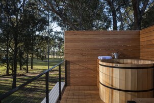 Una sauna de madera con un banco, rodeada por una terraza de madera y una barandilla, ubicada en una zona boscosa.