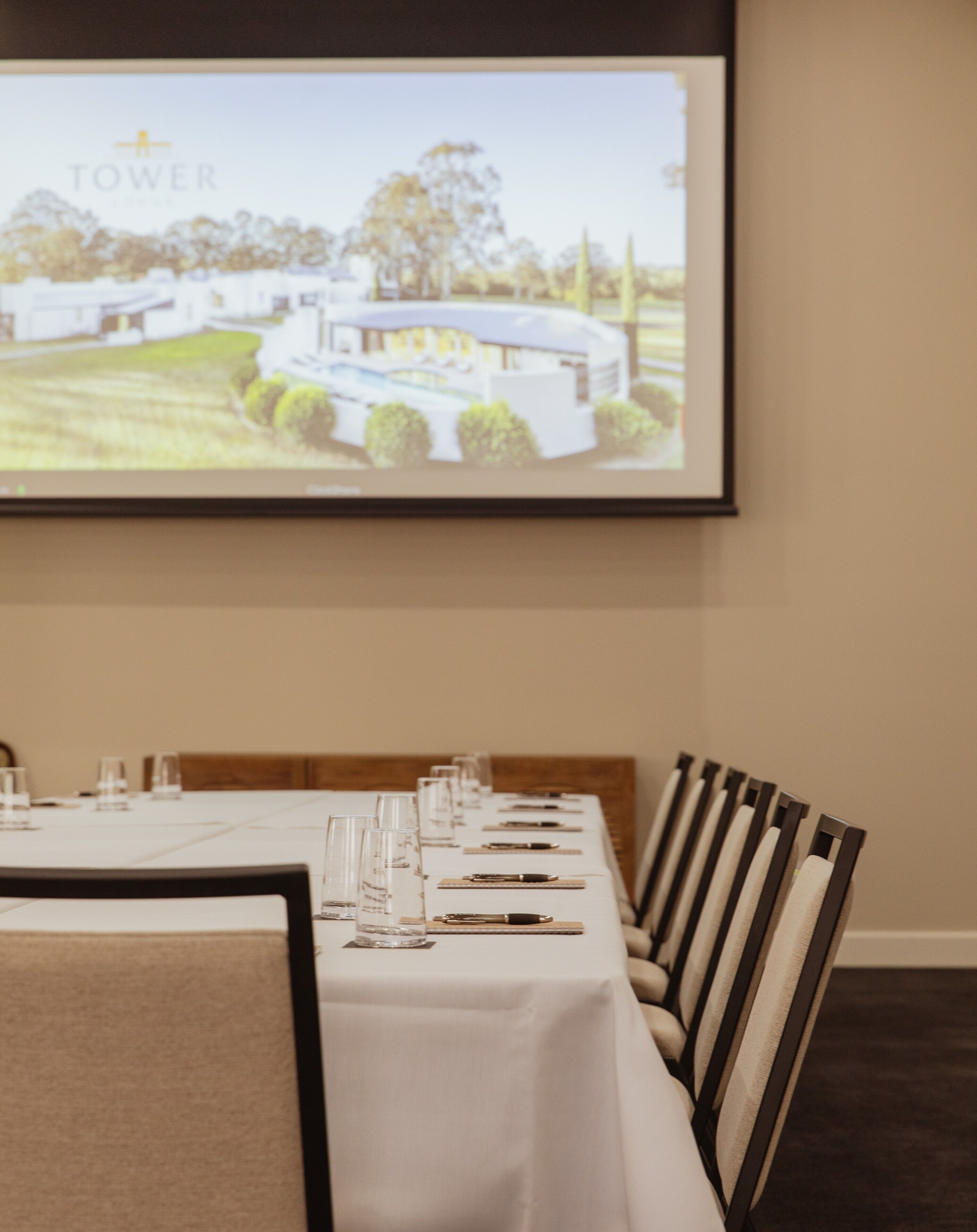 Un salón de conferencias con una larga mesa dispuesta para una reunión, una pantalla de proyector mostrando una imagen de un edificio y una pared color beige.
