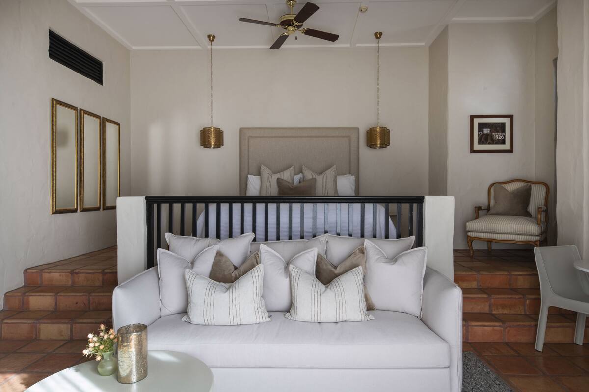 A modern living room with a white sofa, a large bed with a headboard, and a ceiling fan. There are two hanging lamps and a framed picture on the wall.