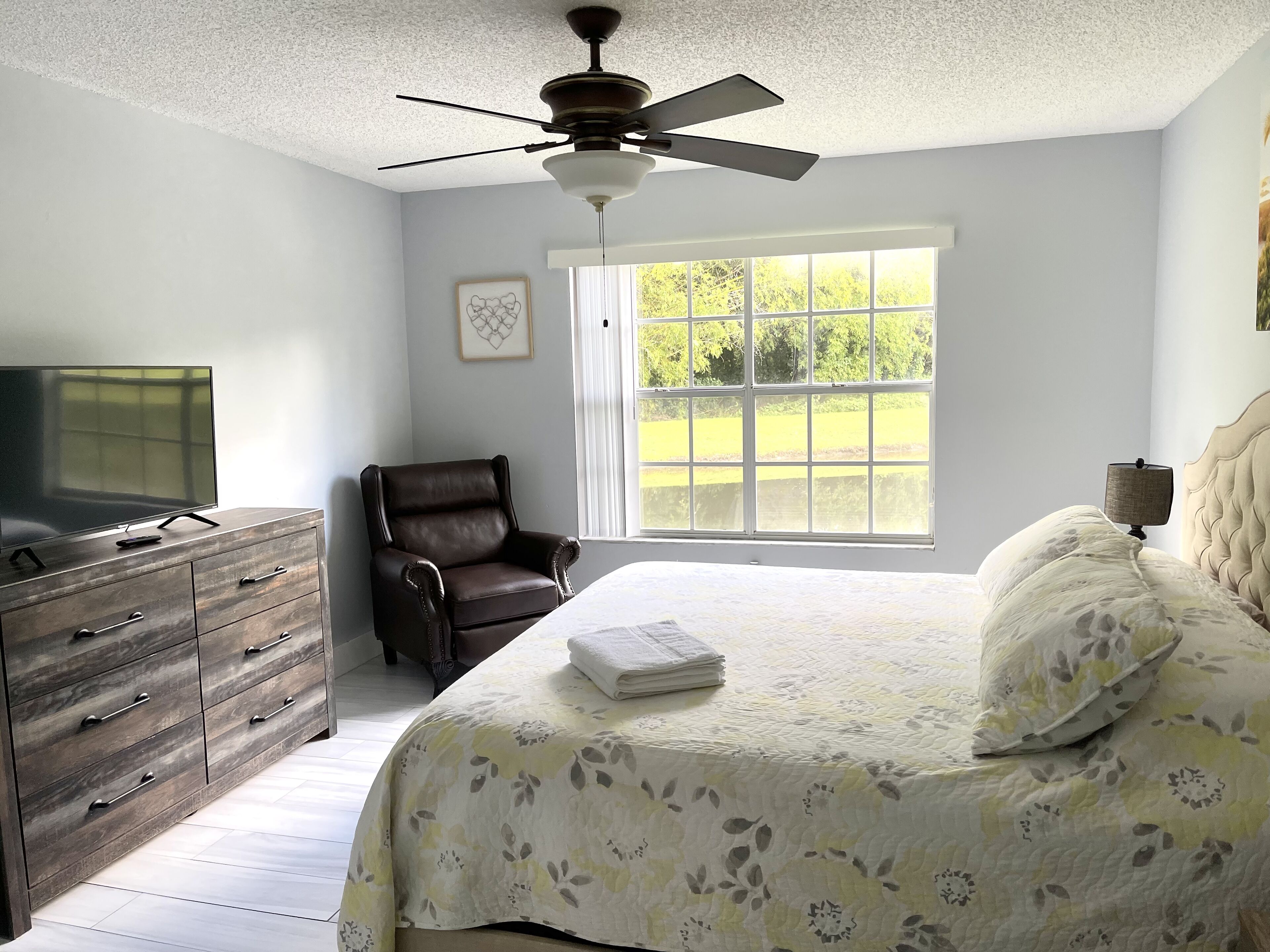 Un dormitorio con cama, una silla, cómoda, ventilador de techo y ventana con vista a follaje.