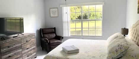 Un dormitorio con cama, una silla, cómoda, ventilador de techo y ventana con vista a follaje.