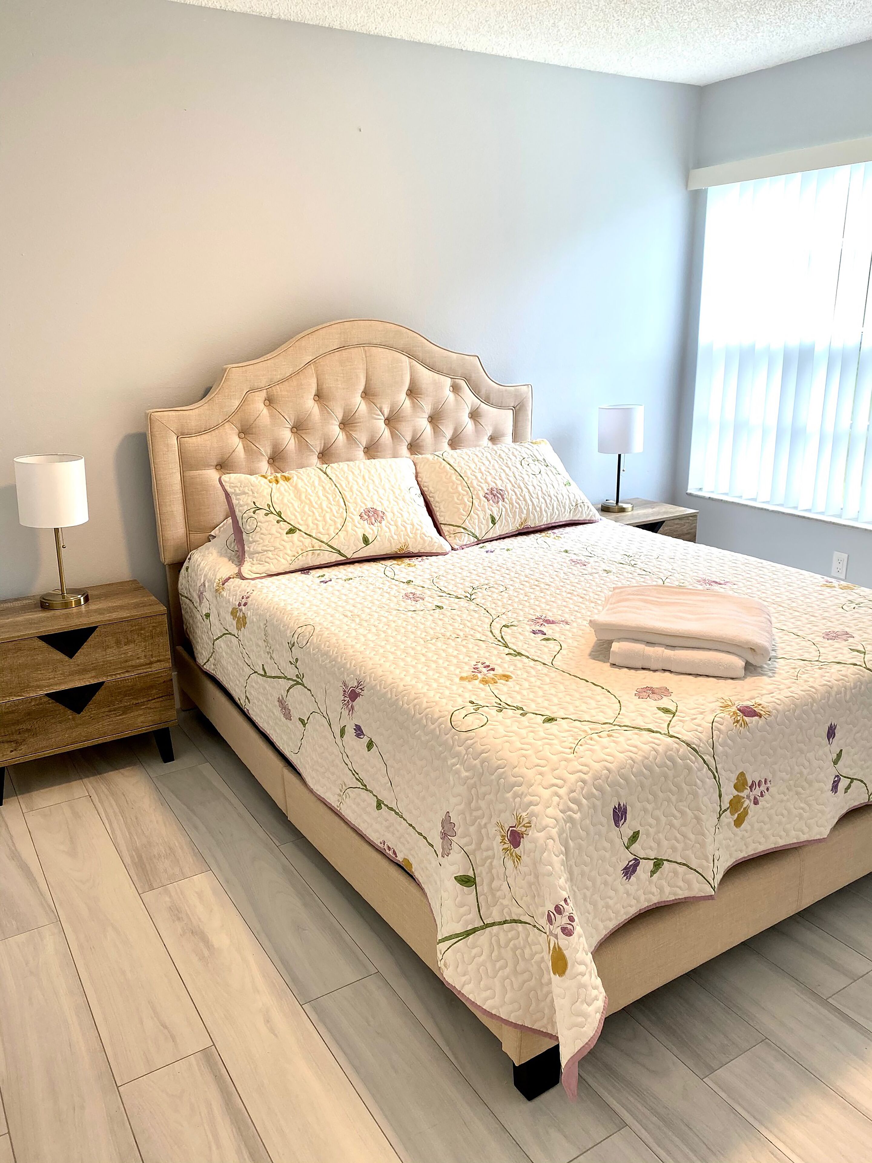 Un dormitorio con una cama grande, una mesita de noche, una lámpara y una ventana con persianas.